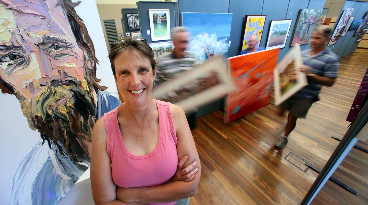 Moira Kirkwood, in front of a portrait painted by Ahn Do, as Frank Nowlan, Ron Buchan and Brian Cummins are busy hanging art works at the Thirroul Library and Community Centre. Picture: KIRK GILMOUR